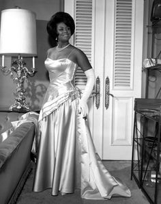 an old photo of a woman in a dress standing next to a bed and lamp