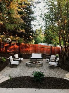 an outdoor fire pit surrounded by chairs and trees with string lights on the fence behind it