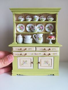 a hand is holding a doll's toy in front of a green china cabinet