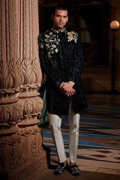 a man standing next to a pillar wearing a black jacket and white pants with flowers on it