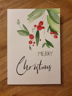 a merry christmas card with watercolor leaves and berries on it, sitting on a wooden surface