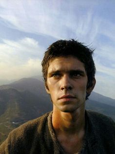 a young man standing on top of a mountain looking at the camera with mountains in the background