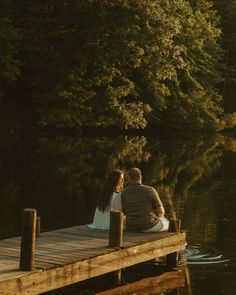 a man and woman sitting on a dock next to the water with trees in the background