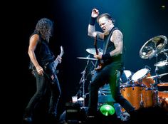 two men on stage with guitars and drums in front of them, one man is holding his arm up