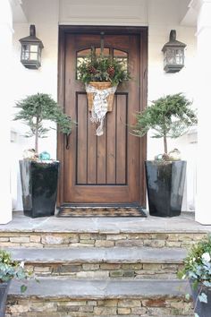 two potted trees are on the front steps next to a wooden door and light fixture