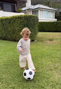 a young boy kicking a soccer ball on the grass in front of a house with hedges