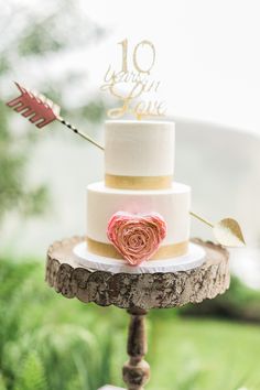 a three tiered wedding cake with an arrow and rose decoration on the top is sitting on a tree stump