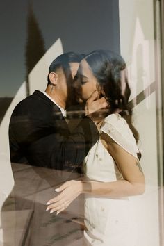 a man and woman are kissing in front of a glass window with the reflection of trees behind them