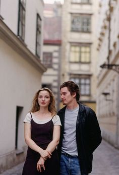 a man and woman standing in an alleyway next to each other with buildings behind them