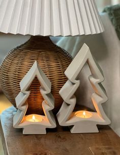 two white candles sitting on top of a wooden table next to a basket and lamp