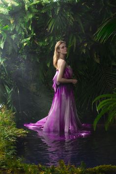 a woman in a purple dress is standing in the water near some plants and trees