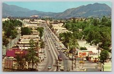 an old photo of a city street with mountains in the backgrouund and cars driving down it