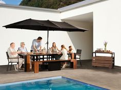 a group of people sitting around a table under an umbrella next to a swimming pool