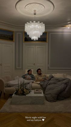 two people sitting on a couch in the middle of a living room with chandelier