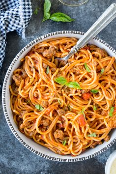 a white bowl filled with spaghetti and meat on top of a blue table cloth next to two silver spoons