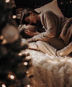 a man and woman laying on a bed in front of a christmas tree with lights