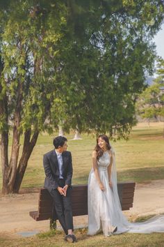 a man and woman are sitting on a bench in front of a tree, looking at each other