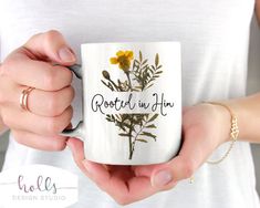 a woman holding a coffee mug with flowers on it that says, rooted in him