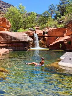 a person swimming in a pool surrounded by red rocks and water falls with trees on either side