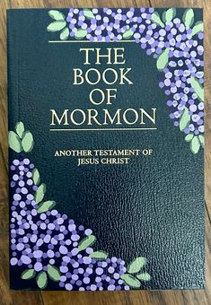 the book of mormon on a wooden table with purple and green flowers in it's center