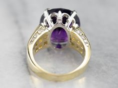 This stunning amethyst cocktail ring has a deep purple amethyst in the center, that really flashes and flickers with vibrant violet reflections! Two rows of square-cut diamonds add sparkle, while round-cut diamonds line the shank of the yellow gold band. Set into a lovely mounting with structurally designed shoulders and a fleur de lis platinum head, this bold statement ring is a beautiful addition to anyone’s collection! Metal: 18K Yellow Gold, Platinum Gem: Amethyst 12.48 Carats Gem Measuremen Fine Jewelry Purple Diamond Ring With Gemstone Accents, Purple Diamond Ring With Gemstone Accents, Formal Purple Diamond Ring With Gemstone Accents, Elegant Purple Diamond Ring With Gemstone Accents, Purple Cushion Cut Brilliant Ring, Purple Brilliant Cushion Cut Rings, Fine Jewelry Purple Rings With Diamond Accents, Luxury Purple Cushion Cut Jewelry, Fine Jewelry Amethyst Diamond Ring