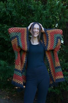 a woman standing in front of some trees holding up a colorful crocheted blanket