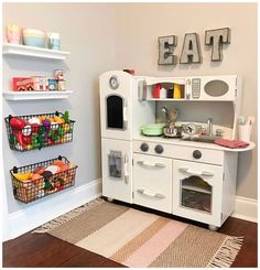 a white play kitchen with lots of food on the shelves and baskets in front of it