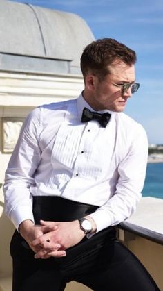 a man in a tuxedo sitting on a ledge next to the ocean wearing sunglasses and a bow tie