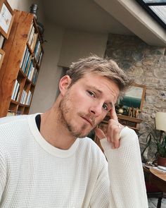 a man sitting in front of a book shelf with his hand on his chin and looking at the camera