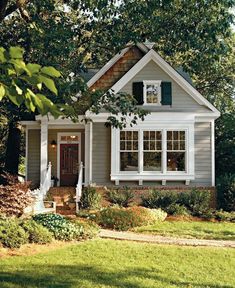 a small gray house with white trim and green shutters on the front door is surrounded by trees