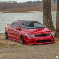 a red car parked in front of some trees near the water with its hood up