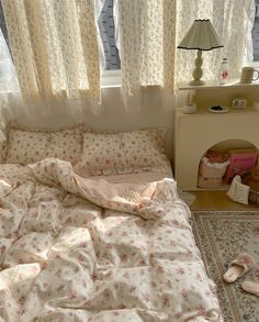 an unmade bed sitting in front of a window next to a rug and lamp