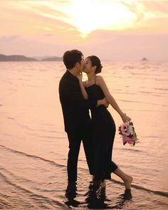 a man and woman standing in the water at sunset kissing each other while holding bouquets