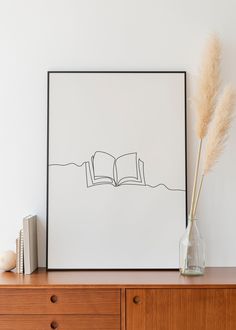 an open book sitting on top of a wooden dresser next to a vase filled with dry grass