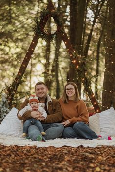 a man and woman sitting on a blanket in the woods with their baby boy, who is wearing an orange hat