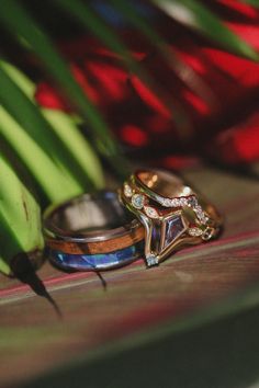 A tropical-themed wedding ring set from Staghead Designs sits beneath palm fronds & bananas. On the left is our "Flynn" titanium wedding band with whiskey barrel oak & paua shell inlays & an elk antler lining. On the right sits our "Bianca" three piece wedding ring set in 14K yellow gold with a kite cut lab-grown alexandrite engagement ring with diamond & alexandrite accents on a scallop-design band paired with two diamond & alexandrite accented tracers. Whiskey Barrel Wedding Ring, Antler Wedding Rings, Matching Wedding Ring Sets, Beachy Wedding, Handcrafted Engagement Ring, Elk Antler, Wood Wedding Ring, Matching Wedding Rings, Staghead Designs