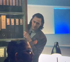 a man is standing in front of a book shelf with papers on it and looking at another man's face