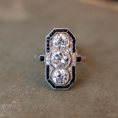 This antique Art Deco cocktail ring features 3.37 carats of Old European cut diamonds set in platinum and surrounded by pieces of square cut onyx. The three diamonds sit north-south along the finger in milgrain adorned bezel settings that appear to be floating inside their onyx frames. This ring is a great example of the fabulous angular style of the Art Deco era and is a perfect piece to accompany you to any cocktail party. Composition: Platinum Era: Art Deco 1915 - 1939 Current Ring Size: 8.50 Vintage Cocktail Rings Art Deco, Art Deco Cocktail Ring, Art Deco Three Stone Diamond White Ring, Art Deco Diamond Three Stone Ring, Diamond White Three Stone Art Deco Ring, Art Deco Three Stone Platinum Jewelry, Classic Wedding Rings With Black Diamonds, Art Deco Jewelry With Center Stone, Classic Rings With Black Diamonds And Cubic Zirconia