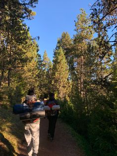 two people walking down a dirt path in the woods with bags on their back and one person carrying an object