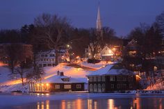 a small town is lit up at night by the water's edge with snow on it