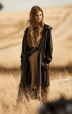 a woman standing in the middle of a field wearing a black jacket and tan dress