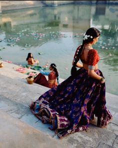 two women in sari sitting on the edge of a body of water with other people around