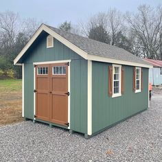 a green shed with brown shutters and two windows on the front, sitting in gravel next to trees