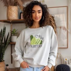 Embrace your green thumb with our Crazy Plant Lady sweatshirt! This shirt features a vibrant design in shades of green that celebrates your love for all things leafy. With playful illustrations of lush plants surrounded by quirky typography, it's perfect for anyone who can't resist adding another plant to their collection. Made from a heavy blend of cotton and polyester, this sweatshirt is both comfortable and warm, making it the ideal addition to any plant lover's wardrobe. Also available as a Green Relaxed Fit Top With Funny Print, Green Long Sleeve Top With Slogan, Green Cotton Sweater With Screen Print, Green Long Sleeve Slogan Top, Green Cotton Sweater With Graphic Print, Summer Crew Neck T-shirt With Plant Print, Green Graphic Tee Cotton Sweatshirt, Green Cotton Sweatshirt With Text Print, Green Long Sleeve T-shirt With Slogan