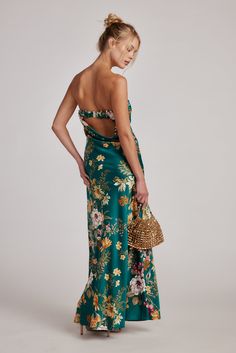 a woman in a green floral print dress with her back to the camera, holding a straw bag