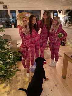 three women in matching pink pajamas standing next to a black dog on the floor near a christmas tree