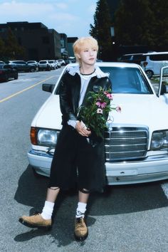 a young man standing next to a white car with flowers in his hand and wearing slippers