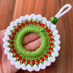 a hand holding a green and red crocheted ornament on top of a wooden table