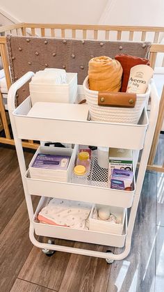 a white cart filled with lots of items on top of a wooden floor next to a baby crib