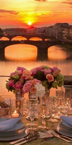 the table is set with flowers and place settings for an elegant dinner in front of a beautiful sunset